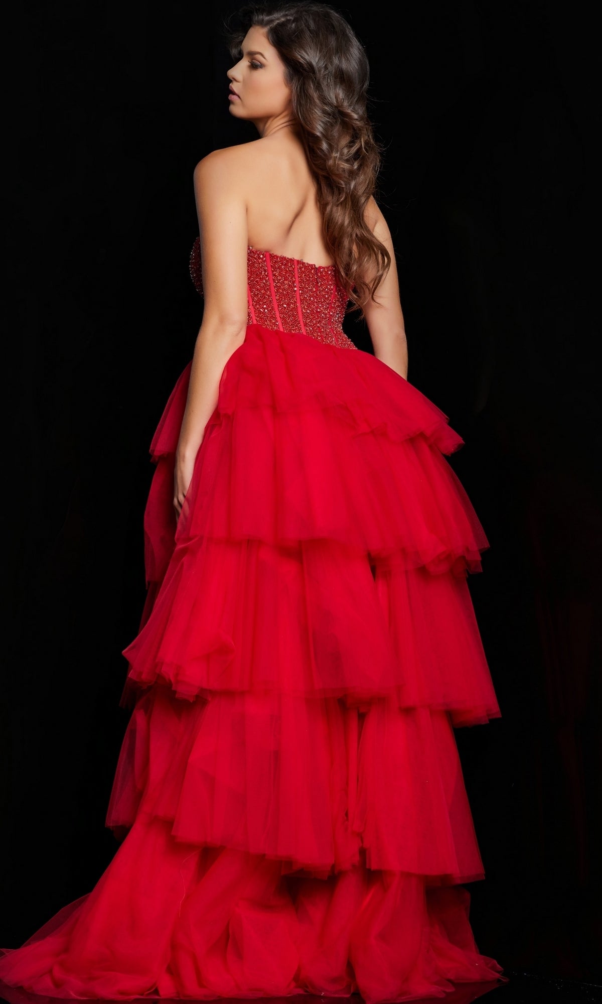 A woman with long wavy hair wears the Long Prom Dress 37210 by Jovani, a strapless tulle ball gown with a layered skirt, standing against a black background and facing away from the camera.